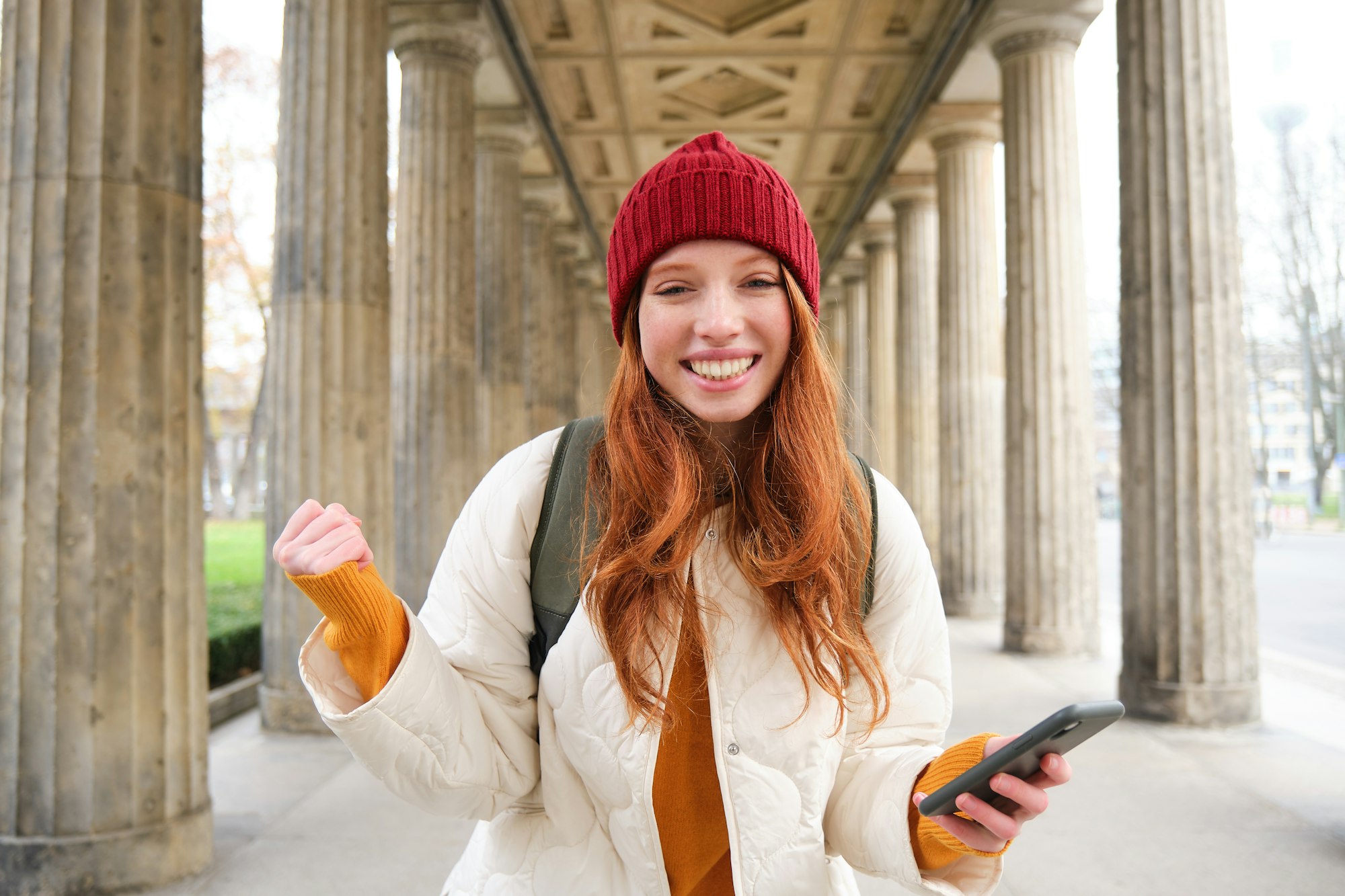 Mobile broadband and people. Smiling redhead 20s girl with backpack, uses smartphone on street